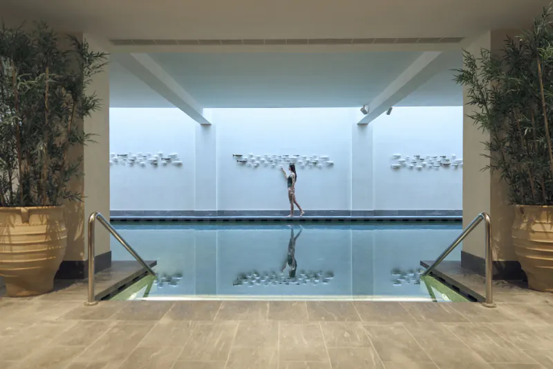 Woman in white swimsuit walking along edge of indoor pool with reflection in calm water, flanked by large potted plants and modern wall art