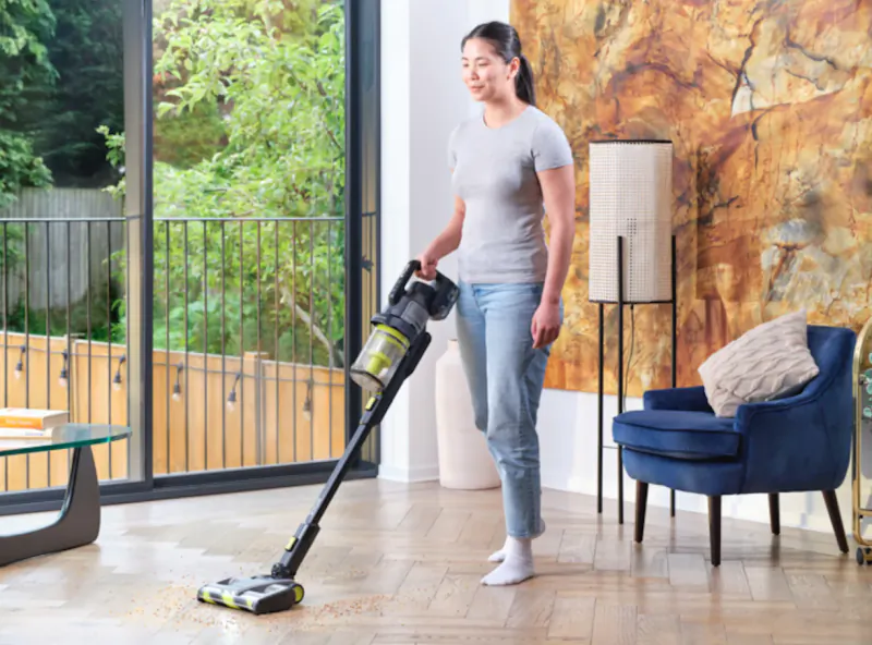 Woman vacuuming crumbs from hardwood floor with a cordless stick vacuum in a bright living room
