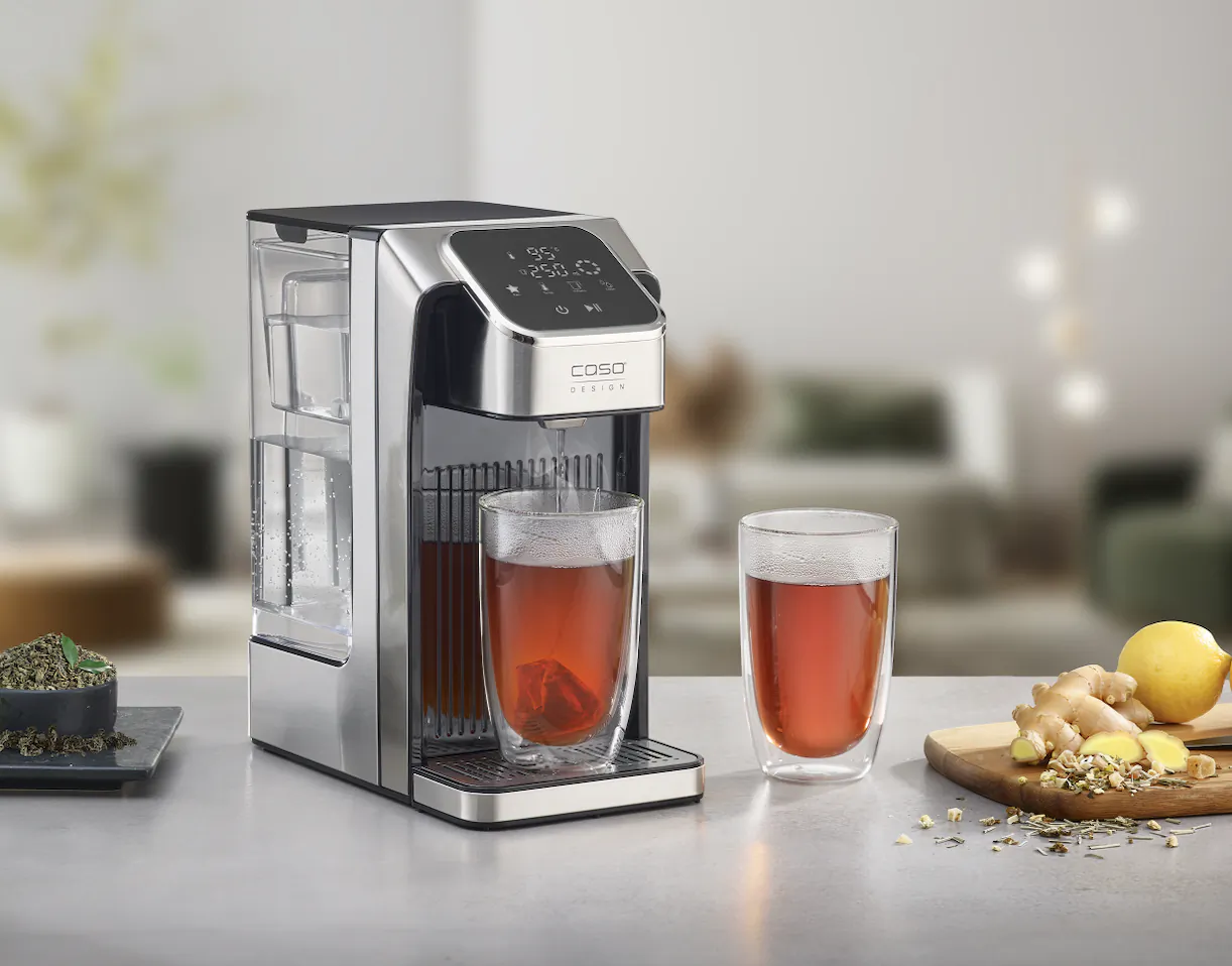 Modern COSORI tea maker dispensing hot tea into a double-walled glass, with another glass and fresh ginger, lemon, and herbs on the counter