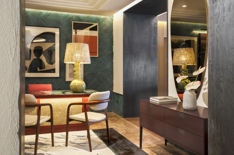 Modern dining area with red and wood table, white chairs, green herringbone wall, abstract art, and tall mirror beside a dark wood console with lamps