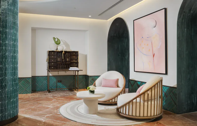 Modern hotel lounge with two white curved armchairs, round coffee table on a circular rug, abstract pink wall art, and a vintage wooden cabinet with decor