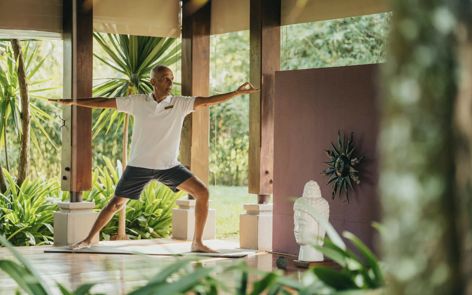 Man in white shirt holding Warrior II yoga pose on mat in open-air pavilion with Buddha statue