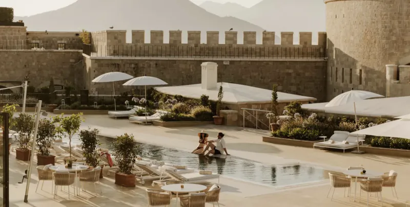 Couple relaxing by the pool at a luxury resort with historic stone fortress and mountains in the background