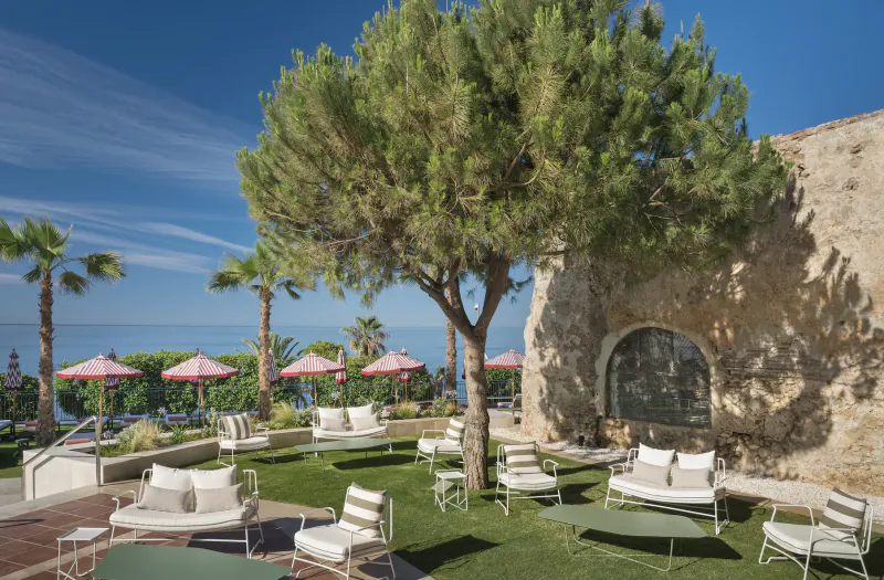 Outdoor lounge area with white chairs and tables on grass beside a stone wall, overlooking the sea with palm trees and red umbrellas