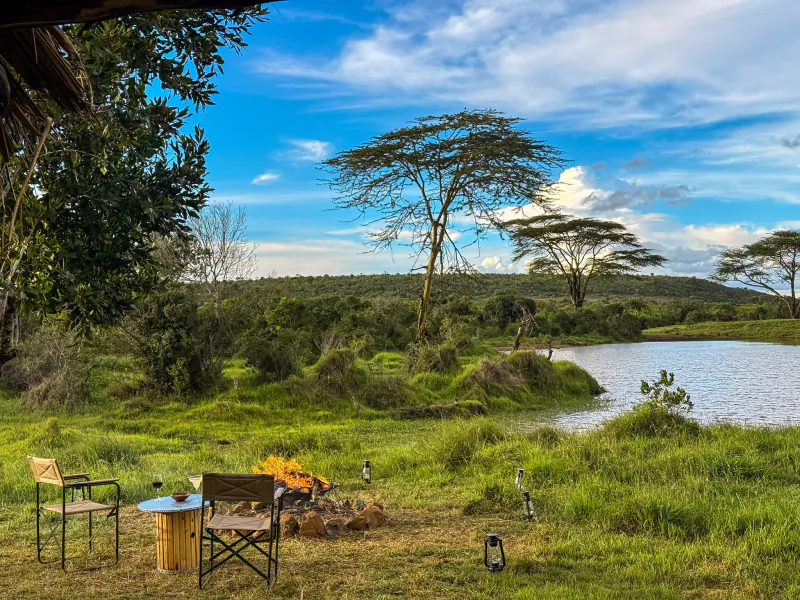 Safari camp setup with two chairs and a table around a campfire overlooking a lake and acacia trees in Laikipia, Kenya