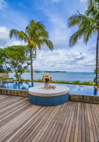 Woman in a straw hat sitting on a round poolside daybed overlooking the ocean with palm trees