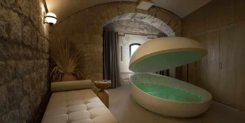 Modern floating pod bathtub with glowing green water inside a rustic stone-walled spa room with a white bench