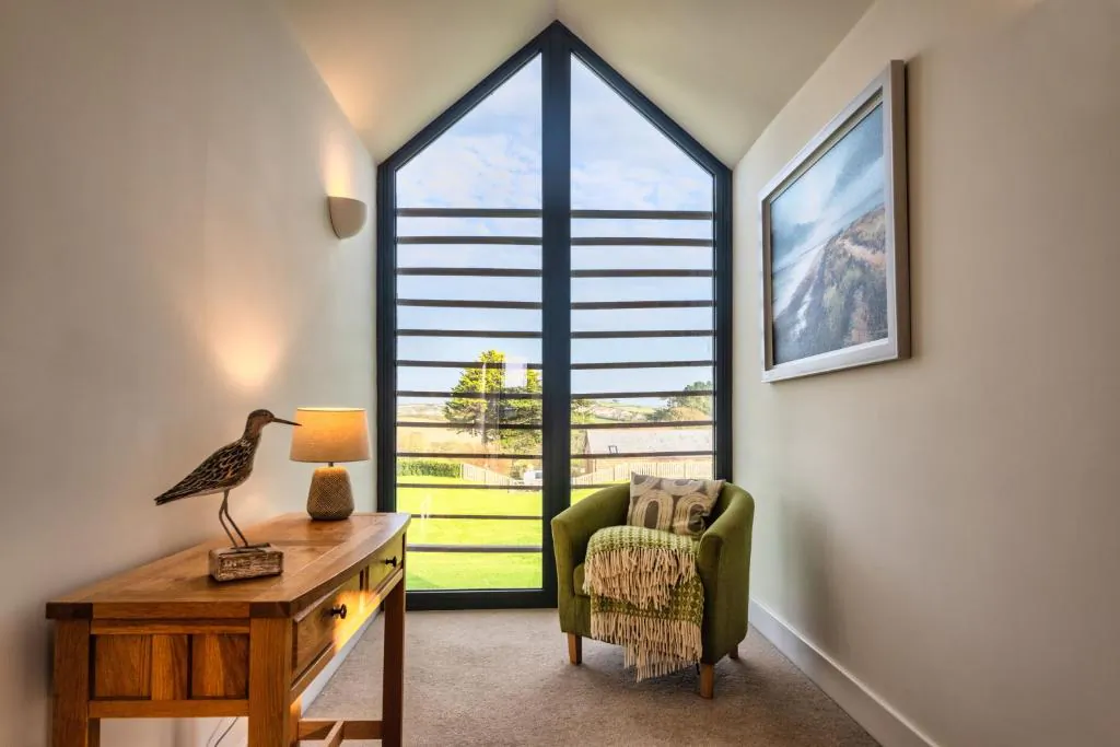 Bright room with tall gabled window overlooking green fields, wooden desk with bird lamp, green armchair, and landscape wall art