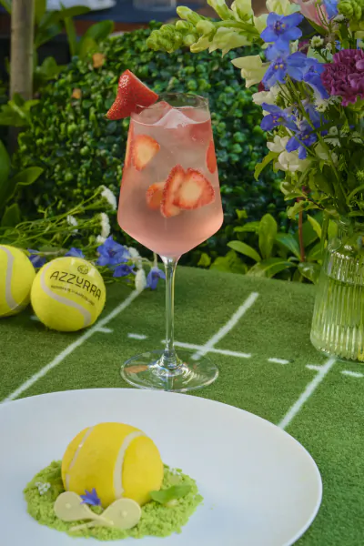 Pink strawberry spritz in a tall glass garnished with fruit slices next to a tennis ball dessert on a white plate, with yellow tennis balls marked Azzurra on artificial grass