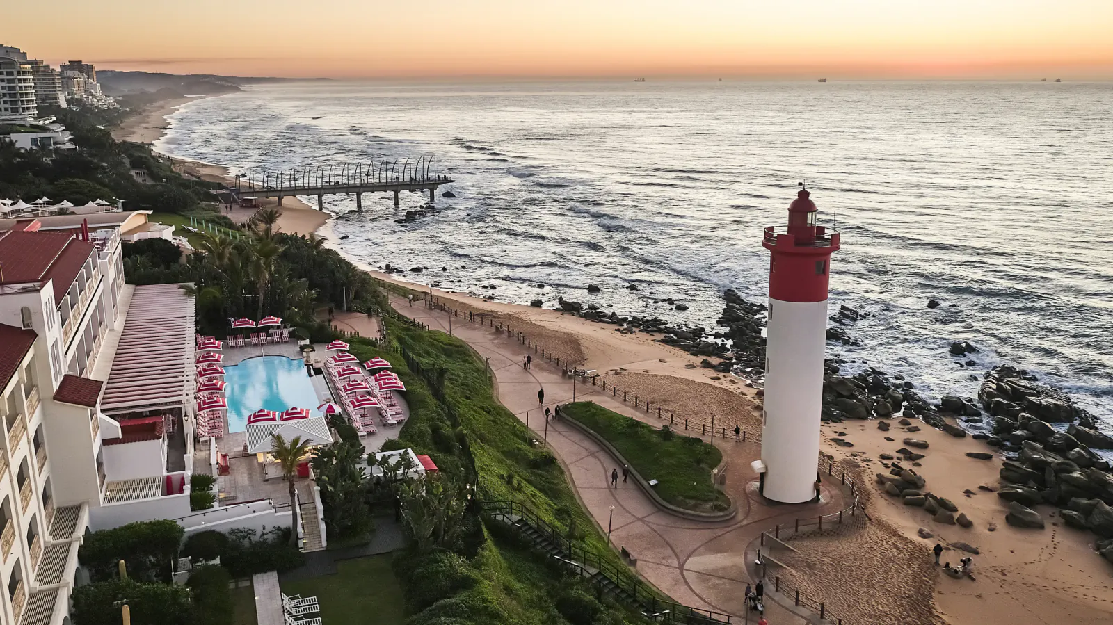 Aerial view of Umhlanga beach at sunset with red and white lighthouse, ocean waves, pier, and resort pool area