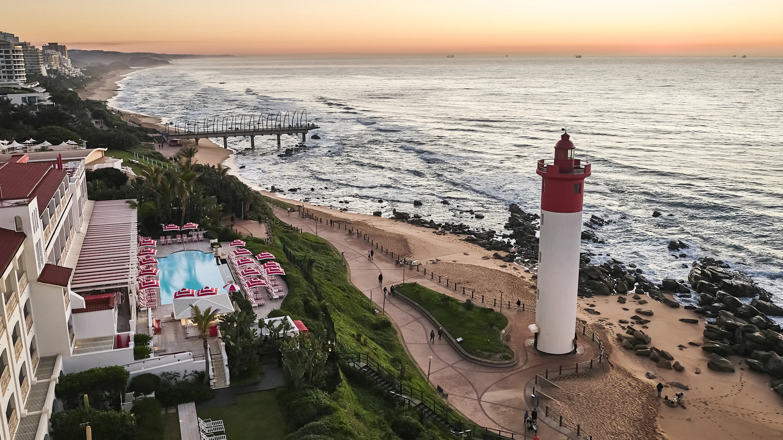 Aerial view of Umhlanga beach at sunset with red and white lighthouse, ocean waves, pier, and resort pool area
