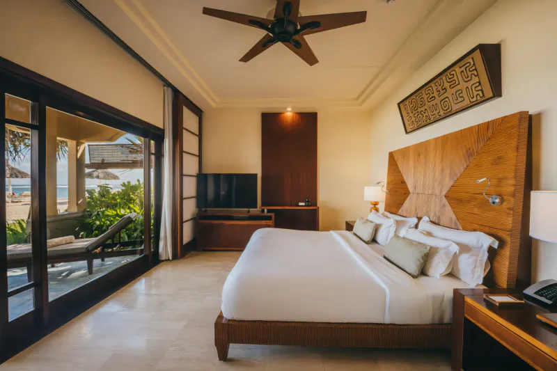 Luxury hotel bedroom with white bed, wood headboard, ceiling fan, and open doors to ocean view balcony