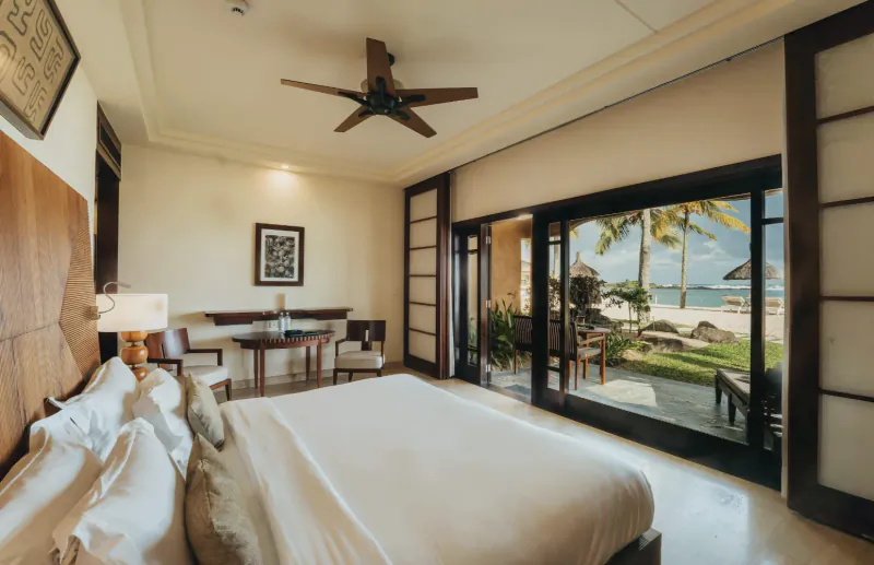 Luxury hotel bedroom with large white bed, open glass doors revealing a tropical beach and ocean view