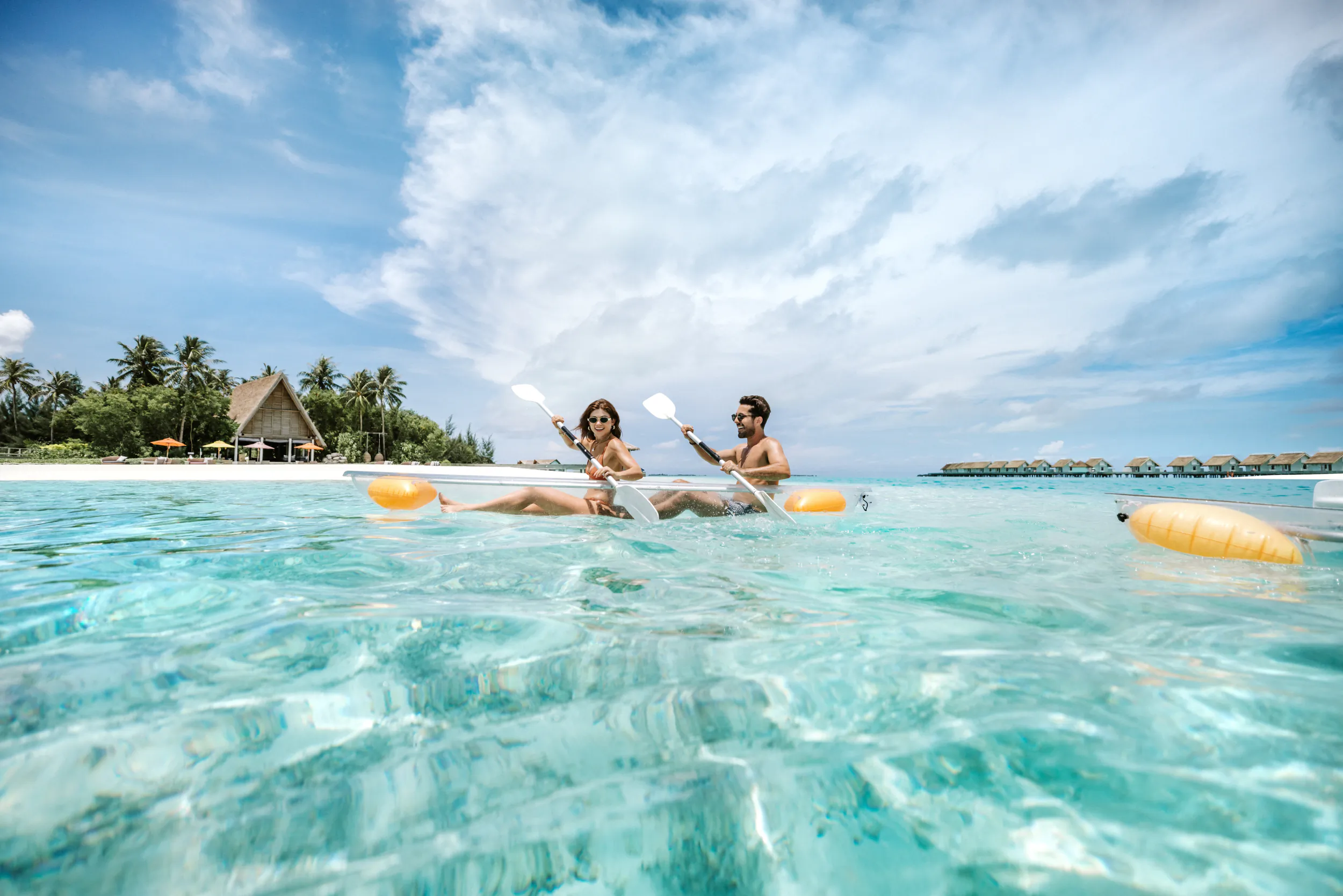 Couple paddling transparent kayaks in clear turquoise water with tropical resort and overwater villas in background