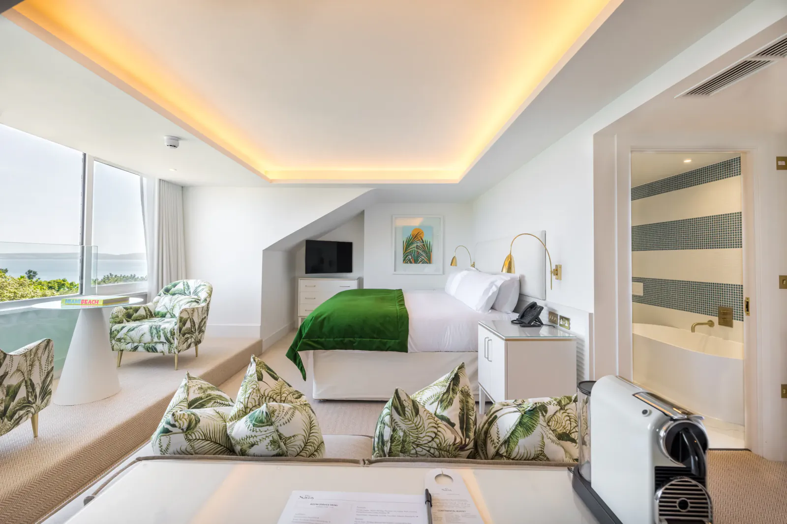 Luxury hotel suite with green bed, patterned chairs, sea-view window, open bathroom, and coffee machine in foreground