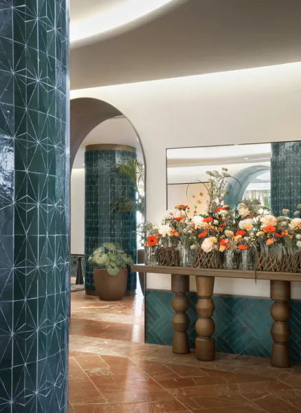 Elegant lobby with teal tiled columns, large floral arrangement on wooden table, terracotta floor, and arched mirrors