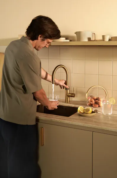 Man in green shirt filling a glass with water from a modern gold kitchen faucet
