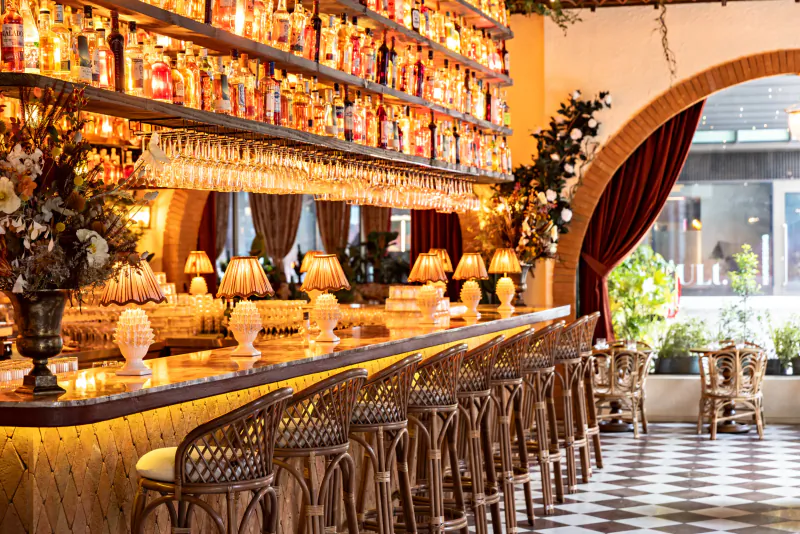 Elegant bar interior with rows of colorful liquor bottles on shelves, woven bar stools along a marble counter, and warm lighting
