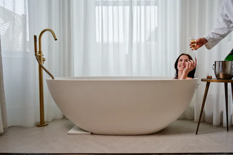 Woman smiling in a white freestanding bathtub as a server offers champagne at The Nici Bournemouth