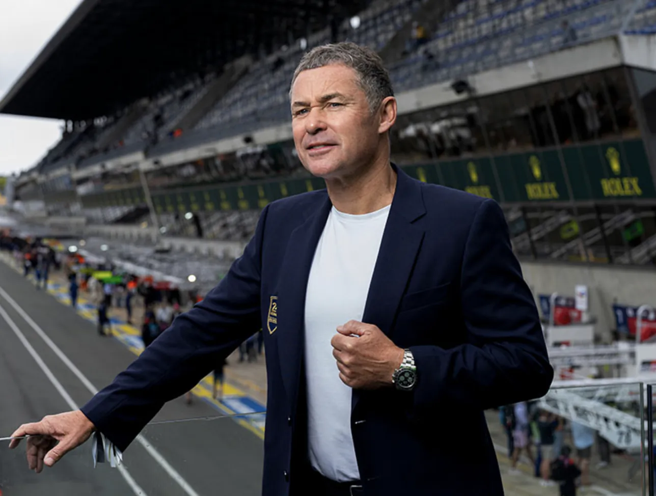 Man in navy blazer gesturing on racetrack with Rolex banners and grandstands behind