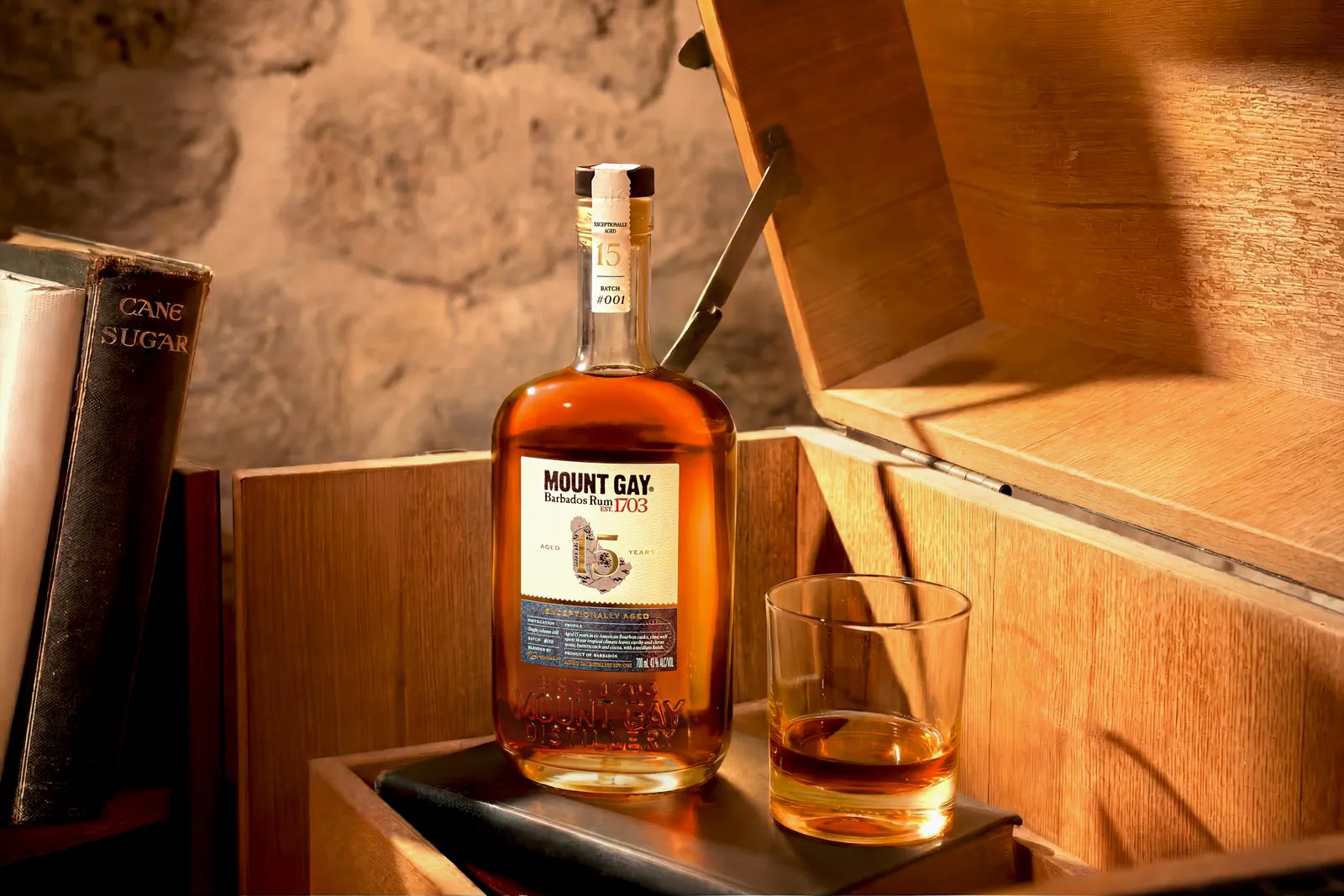 Mount Gay XO rum bottle and glass of amber rum on a wooden chest beside old books