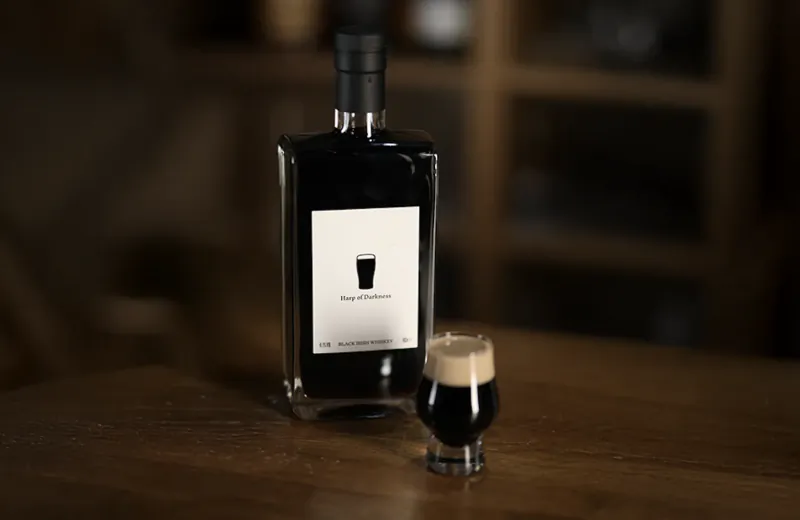 Black whisky bottle labeled 'Hug of Darkness' beside a small glass of dark whisky with foam on a wooden table