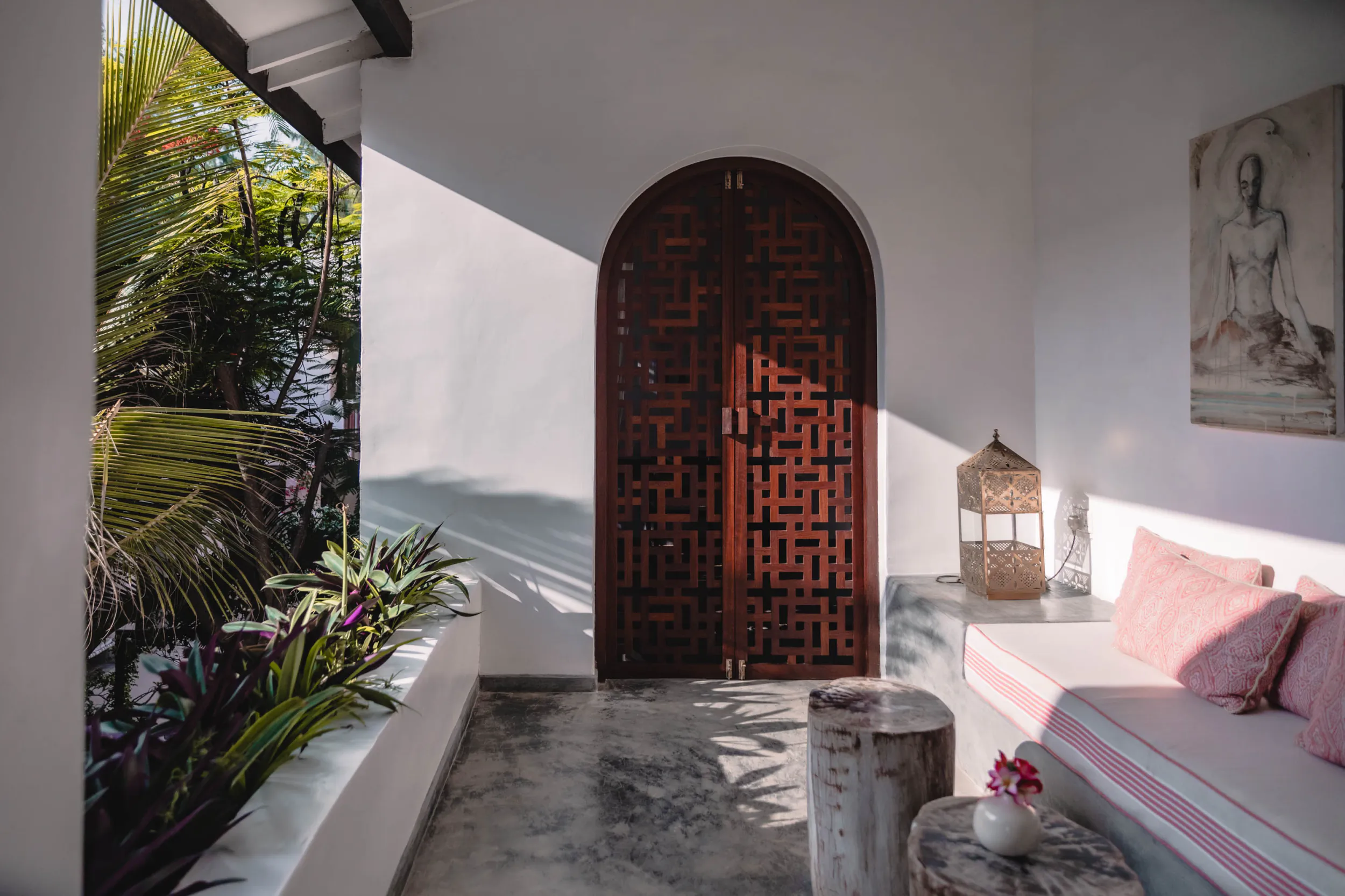 Arched wooden double doors with geometric lattice design in a bright white villa entryway with tropical plants and a daybed