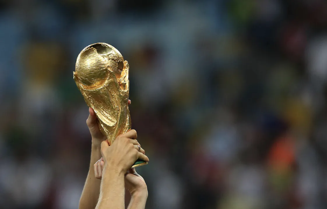 Hands raising the golden FIFA World Cup trophy against a blurred stadium crowd
