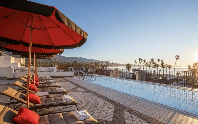 Rooftop pool with red lounge chairs and umbrellas overlooking the ocean at sunset