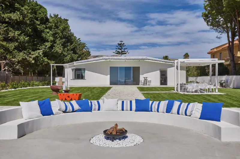 Modern white villa with curved outdoor lounge, blue and white striped cushions, fire pit, and lawn seating area