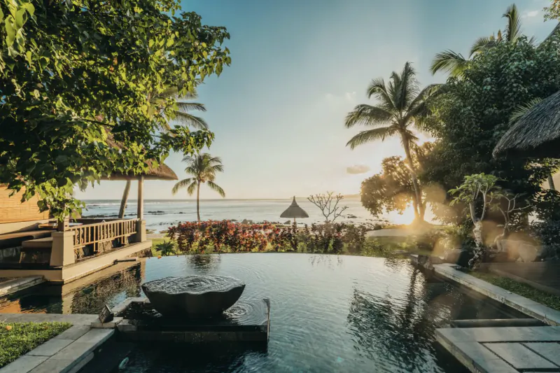Infinity pool overlooking the ocean at sunset with palm trees and thatched-roof villas at Shanti Maurice resort