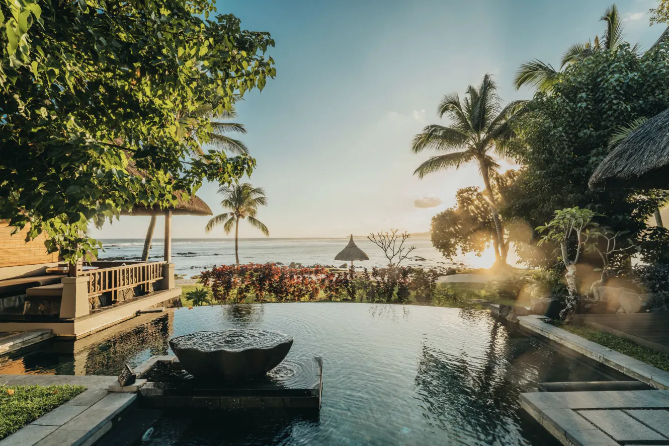 Infinity pool overlooking the ocean at sunset with palm trees and thatched-roof villas at Shanti Maurice resort