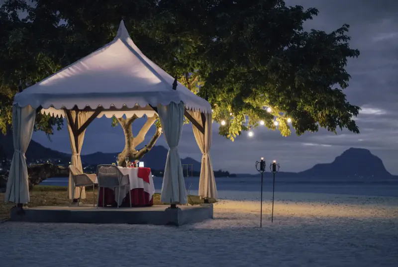 Romantic candlelit dinner setup inside a white canopy tent on a sandy beach at dusk