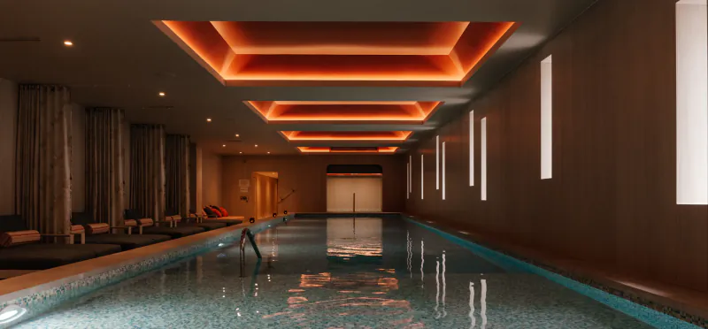 Indoor luxury pool with a person standing in the water, warm orange ceiling lights, lounge chairs, and wooden walls