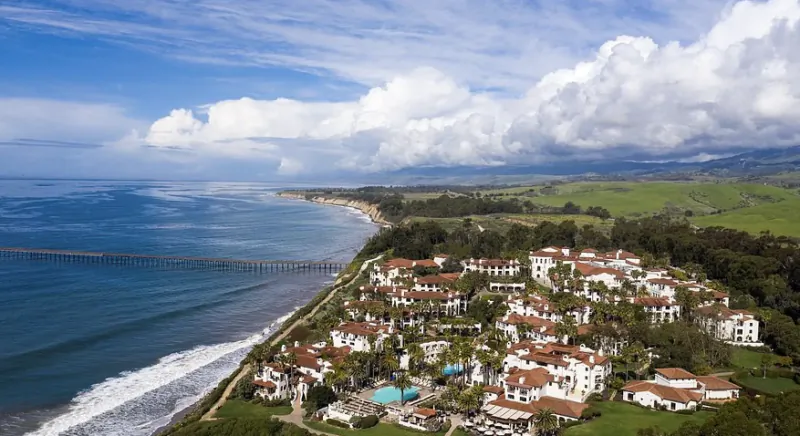 Aerial view of coastal resort with white buildings, red roofs, and a long pier extending into the blue ocean under a cloudy sky
