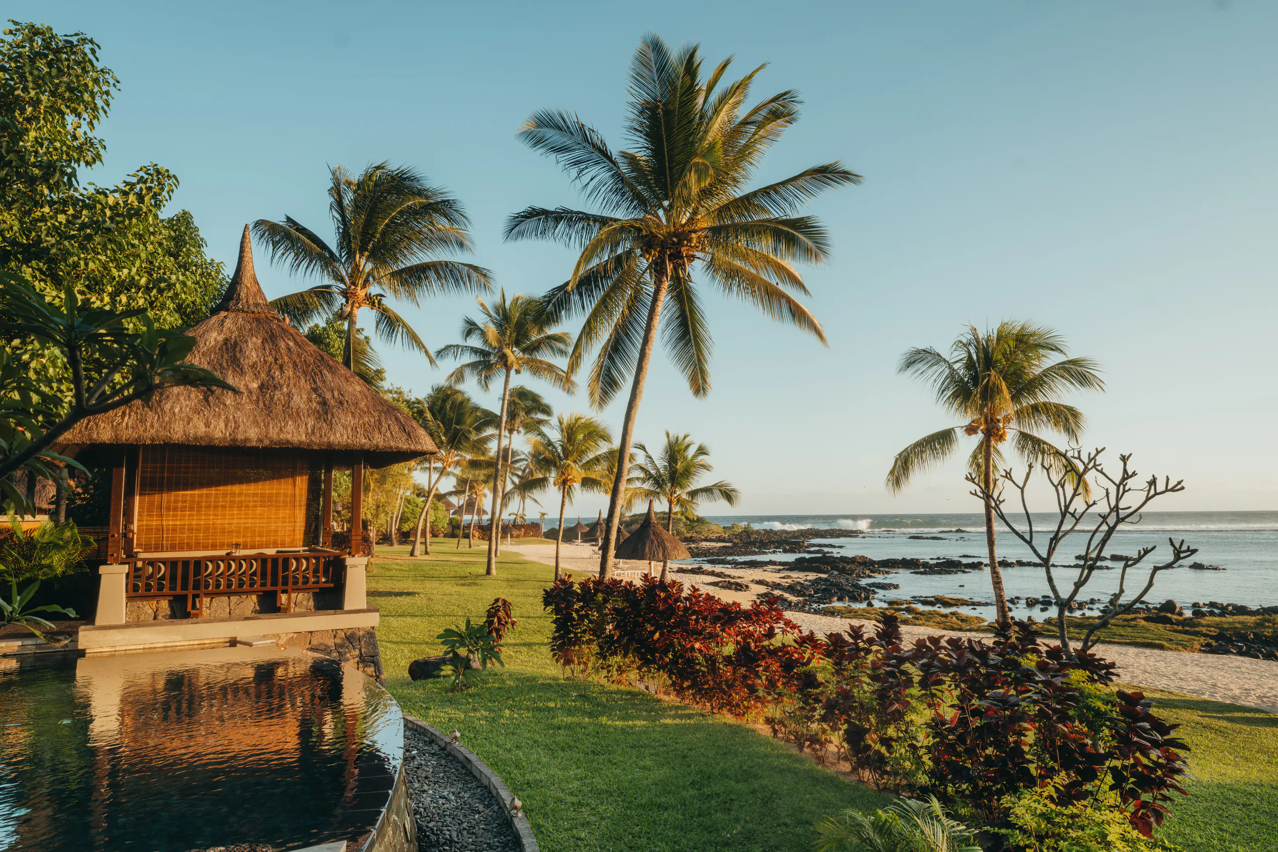 Thatched-roof gazebo beside a reflecting pool overlooking a palm-lined beach and ocean at sunset