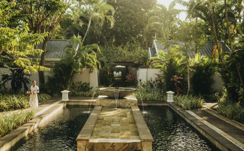 Woman in white dress standing by a tropical resort pool with central fountain and lush greenery