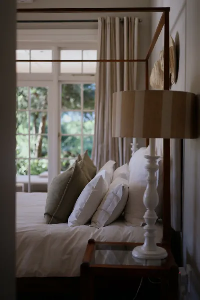 Four-poster bed with white and beige pillows beside a white lamp and nightstand, large window with green view