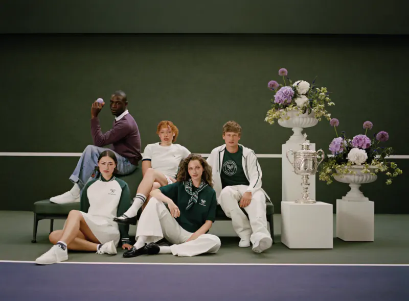 Five models in green and white tennis outfits pose with silver trophies and floral urns on a tennis court