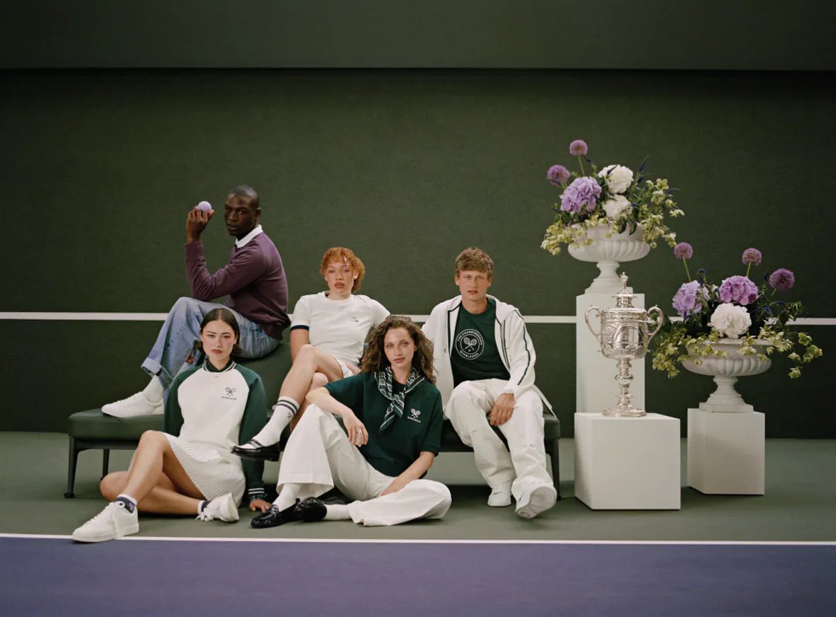 Five models in green and white tennis outfits pose with silver trophies and floral urns on a tennis court