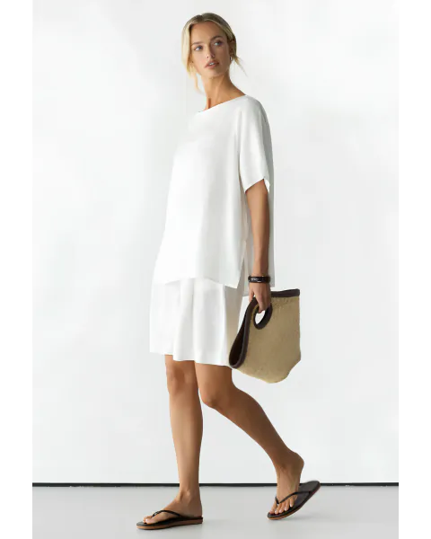 Woman in white satin mini dress holding woven tote bag, walking barefoot in flip-flops against white backdrop