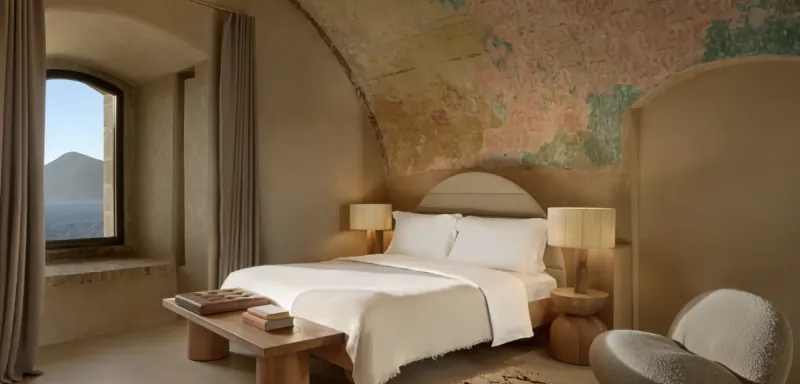 Serene bedroom with a large bed, white bedding, two bedside lamps, wooden bench with books, chair, window overlooking sea and mountains, mural on curved wall