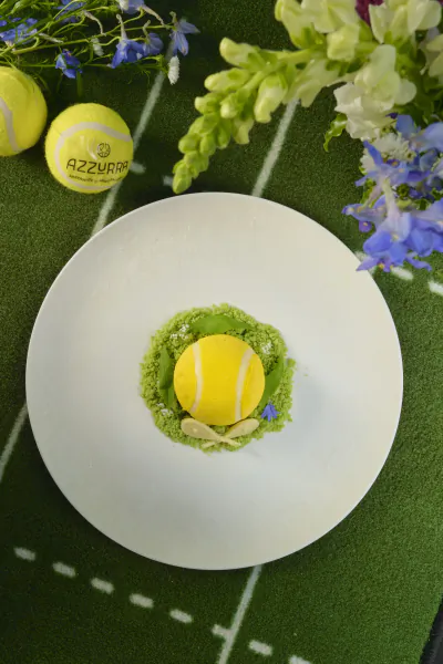 Yellow tennis ball dessert on green matcha base with slivered almonds, surrounded by tennis balls and flowers on artificial grass court