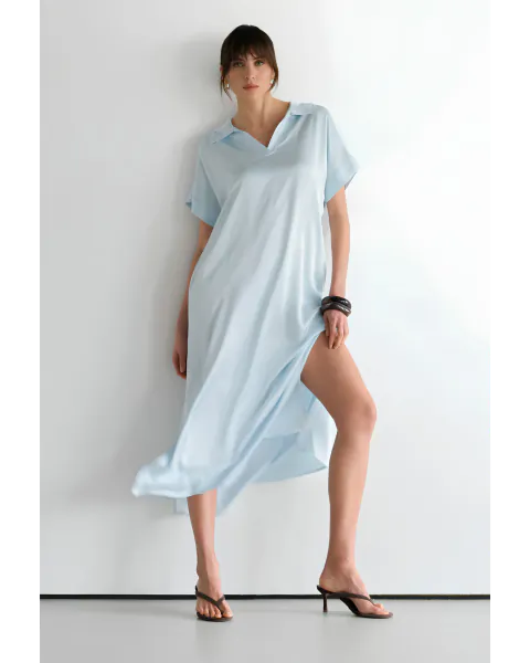 Woman in light blue satin shirt dress posing against white wall with one leg raised