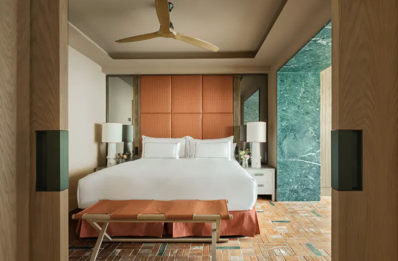 Modern hotel bedroom with large white bed, orange headboard, bench, and green marble wall