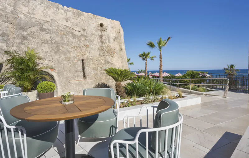 Outdoor terrace with round wooden table and teal chairs overlooking the sea beside a historic stone wall