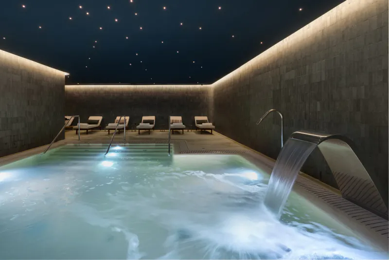 Indoor spa pool with cascading waterfall, lounge chairs along stone walls, and starry night ceiling lights