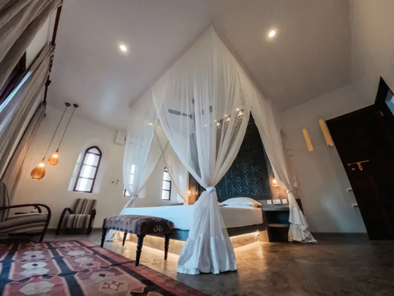 Elegant bedroom with a four-poster bed draped in sheer white canopy curtains, patterned rug, and arched window in a luxury villa
