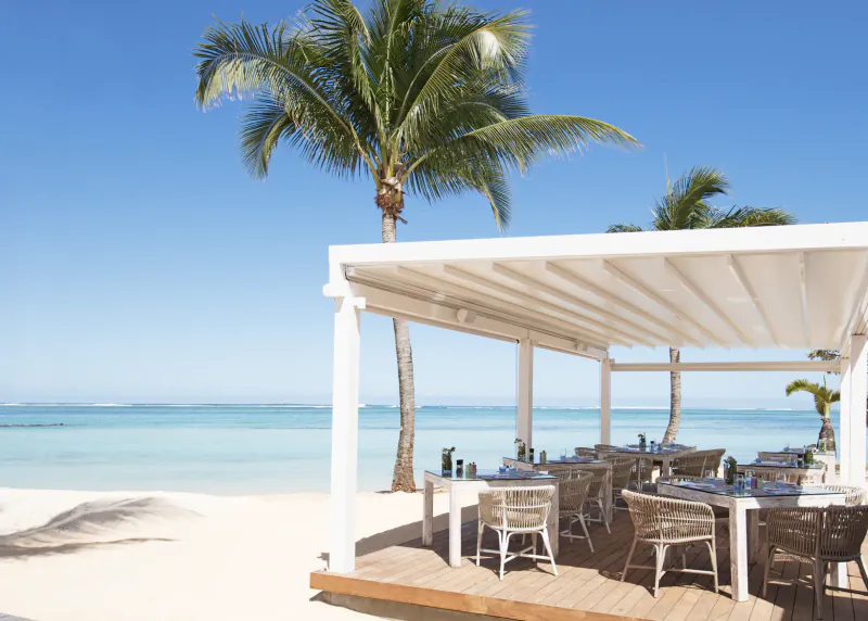Beachfront dining tables under a white pergola with ocean and palm trees in Mauritius