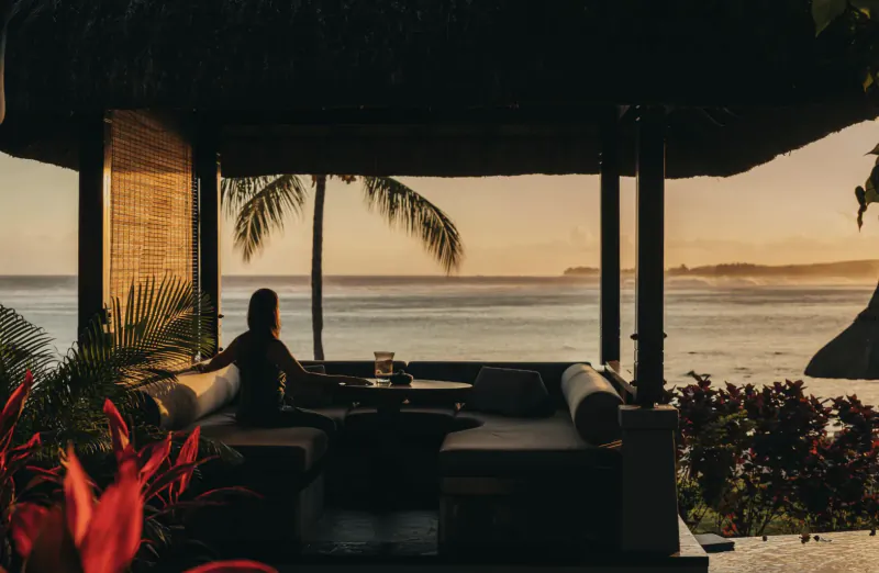 Woman seated on a lounge sofa under a thatched-roof cabana overlooking the ocean at sunset